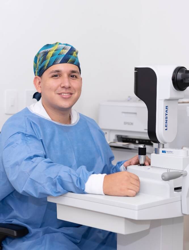 Hay un profesional de la salud sonriendo de frente y a su lado hay un equipo oftalmológico.