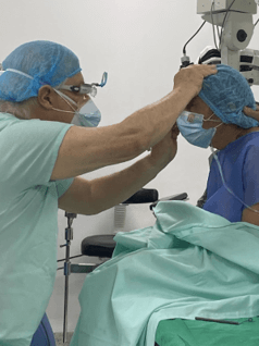nuestros servicios En un ambiente médico hay un paciente sentado con ropa quirúrgica y envuelto en mantas quirúrgicas, al frente de él hay un profesional de la salud revisando sus ojos.