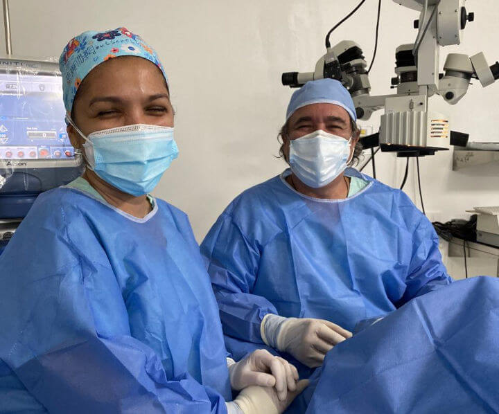En un ambiente de sala de procedimientos hay dos profesionales de la salud, un hombre y una mujer, con ropa quirúrgica y tapabocas, pero se puede ver que sonríen, se nota que hay cubierto un paciente en la mesa, y del techo se ve un equipo que se infiere que es para cirugía oftalmológica.